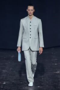 Male model walks the runway in a light gray pinstripe suit with a stand collar, holding a pale blue clutch.