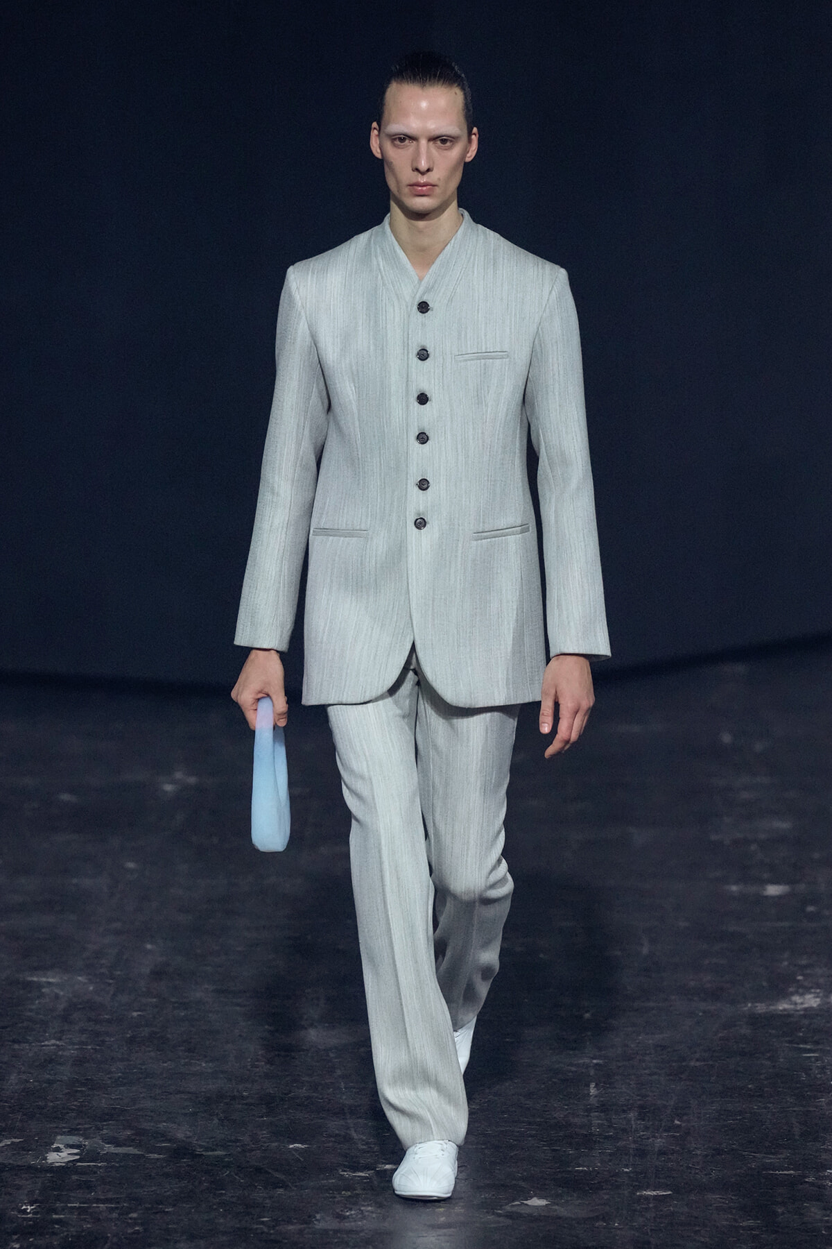 Male model walks the runway in a light gray pinstripe suit with a stand collar, holding a pale blue clutch.