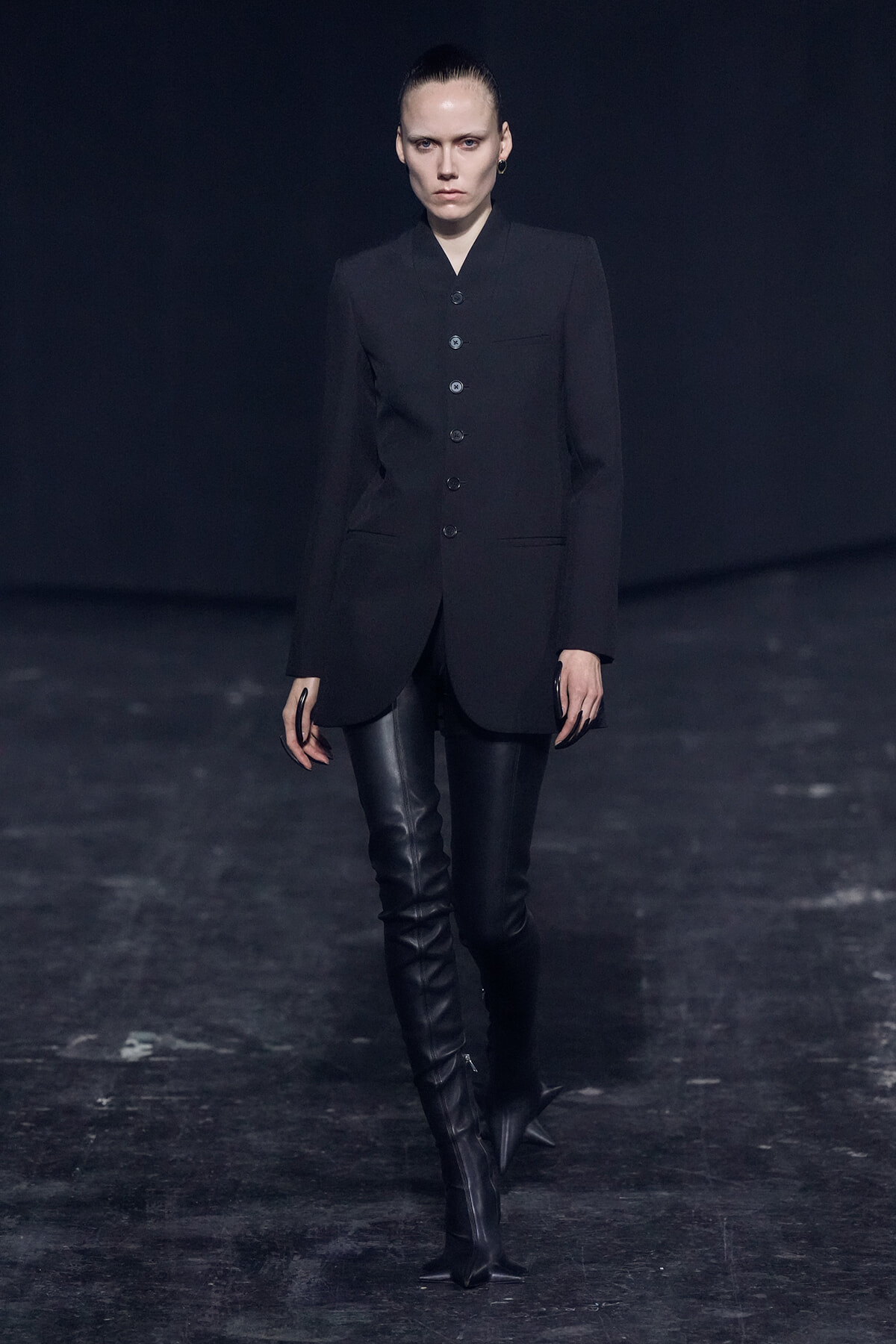 Model walks a runway in a black tailored blazer with large buttons and thigh-high leather boots.