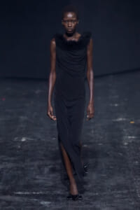 Runway model in a long black dress with a feathered fur collar walking toward the camera.