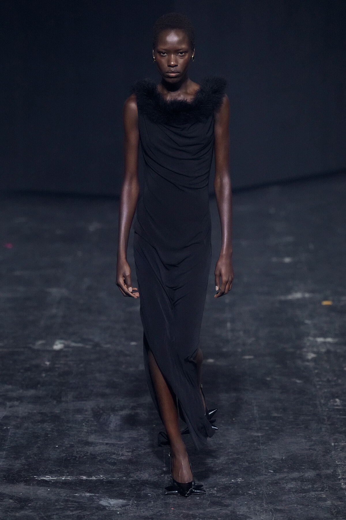 Runway model in a long black dress with a feathered fur collar walking toward the camera.