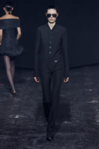 Model wearing a black tailored jumpsuit with a button-front and dark sunglasses walking a runway against a dark backdrop; another model in black dress visible in the background.