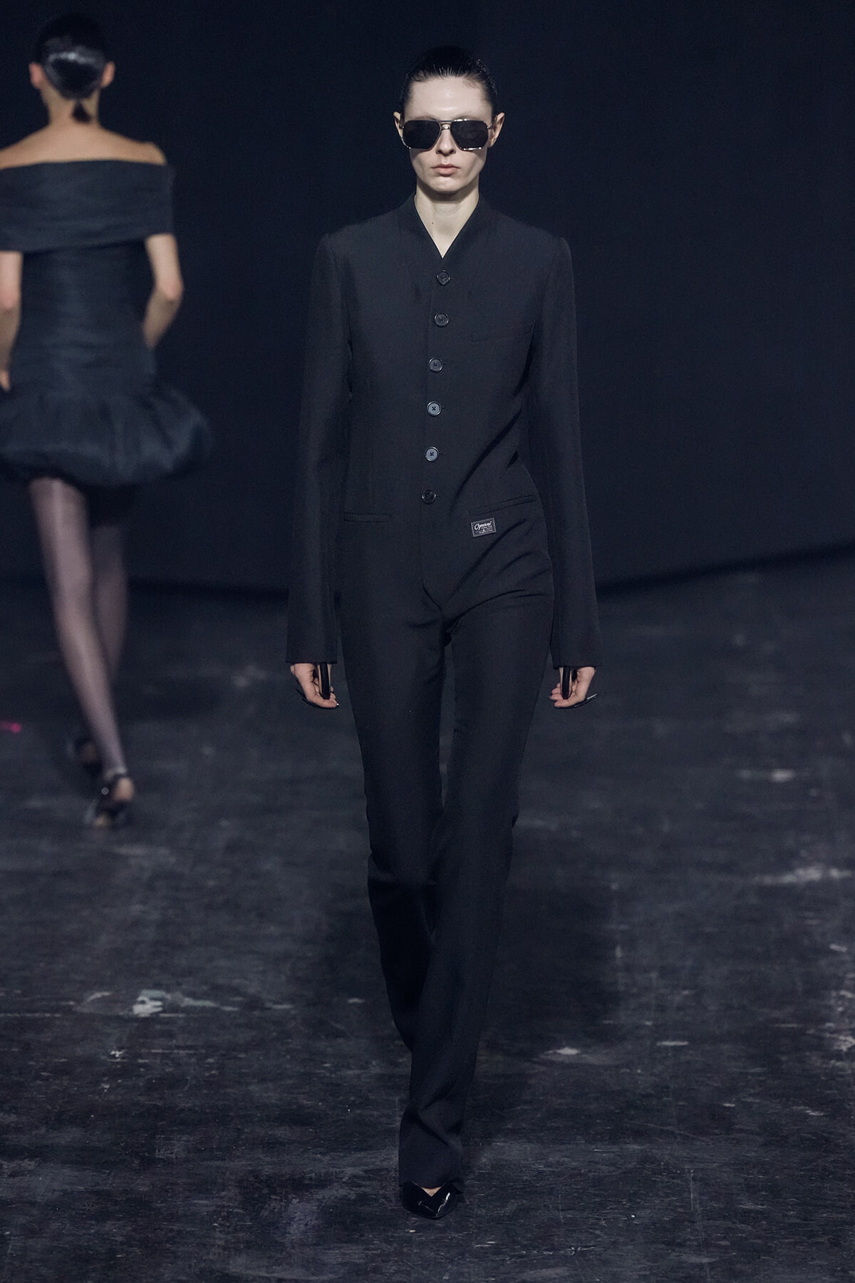 Model wearing a black tailored jumpsuit with a button-front and dark sunglasses walking a runway against a dark backdrop; another model in black dress visible in the background.