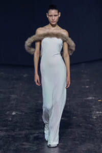Model walking a fashion runway in a white halter gown with a fluffy fur stole draped over her shoulders against a dark background.