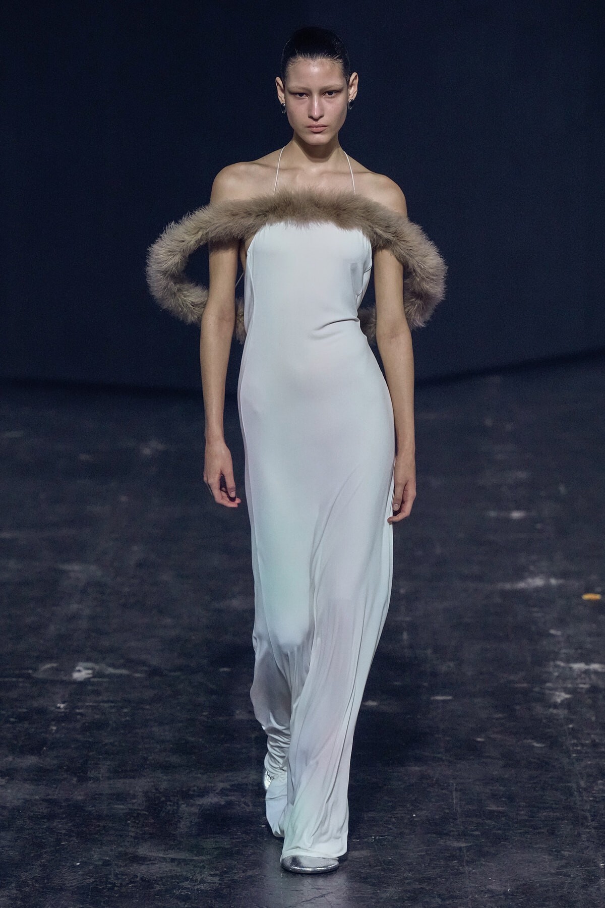Model walking a fashion runway in a white halter gown with a fluffy fur stole draped over her shoulders against a dark background.
