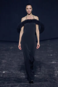 Female runway model wearing a sleek black strapless gown with a thick black fur collar, standing on a dark catwalk with hair slicked back and blue earrings.