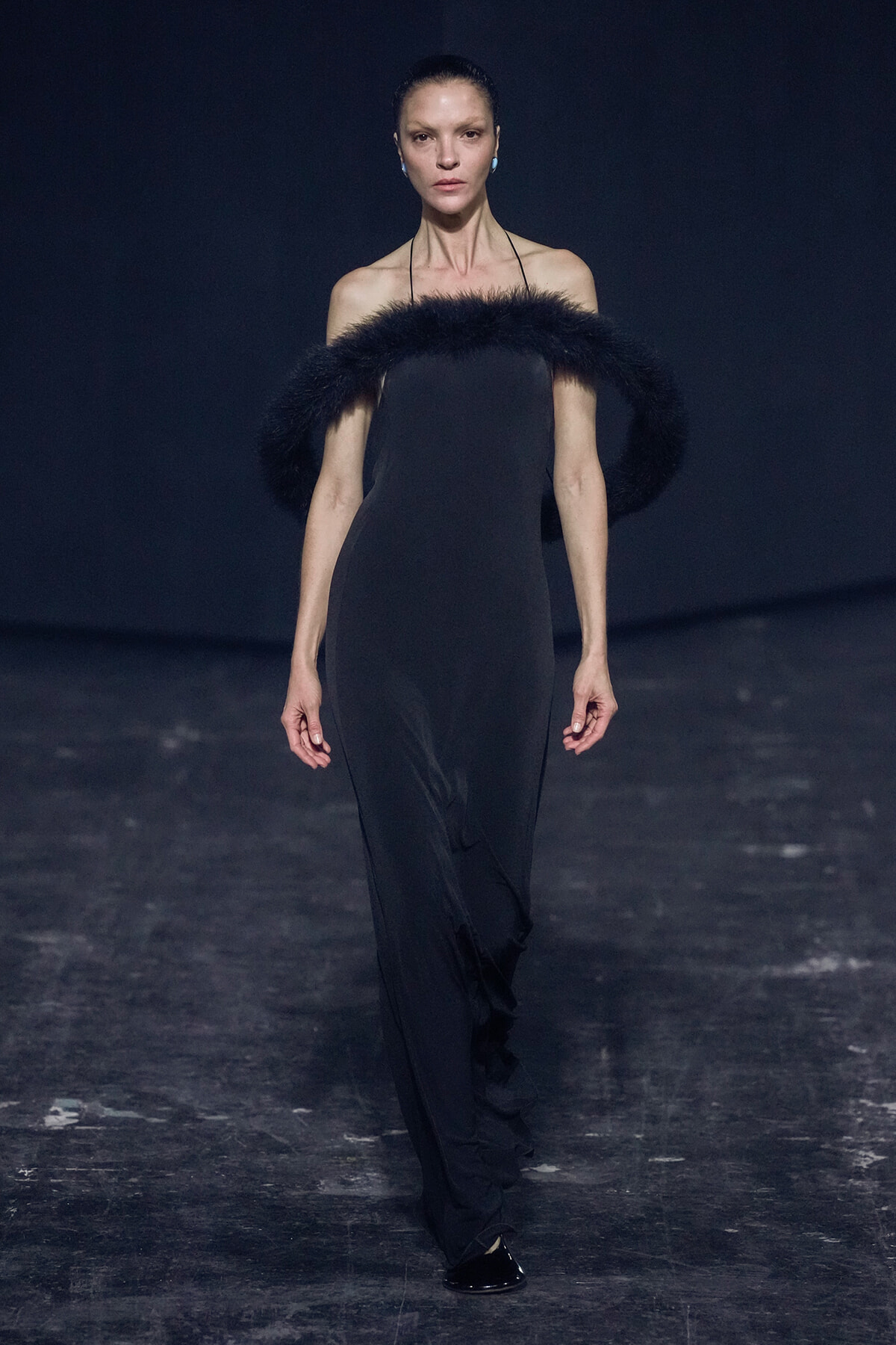 Female runway model wearing a sleek black strapless gown with a thick black fur collar, standing on a dark catwalk with hair slicked back and blue earrings.