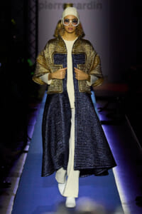 Model walks a blue-lit runway in a metallic gold and navy quilted coat over a white outfit, white beanie and oversized sunglasses, hands adjusting the coat.