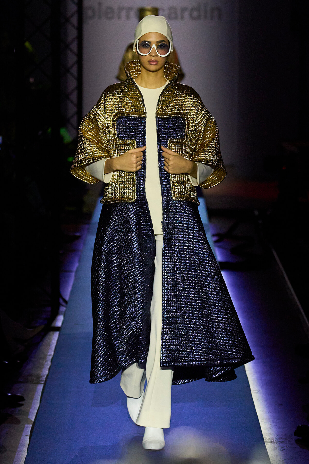Model walks a blue-lit runway in a metallic gold and navy quilted coat over a white outfit, white beanie and oversized sunglasses, hands adjusting the coat.