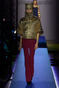 Model walks the runway in a metallic gold textured hooded top with a high neck, paired with burgundy pants.