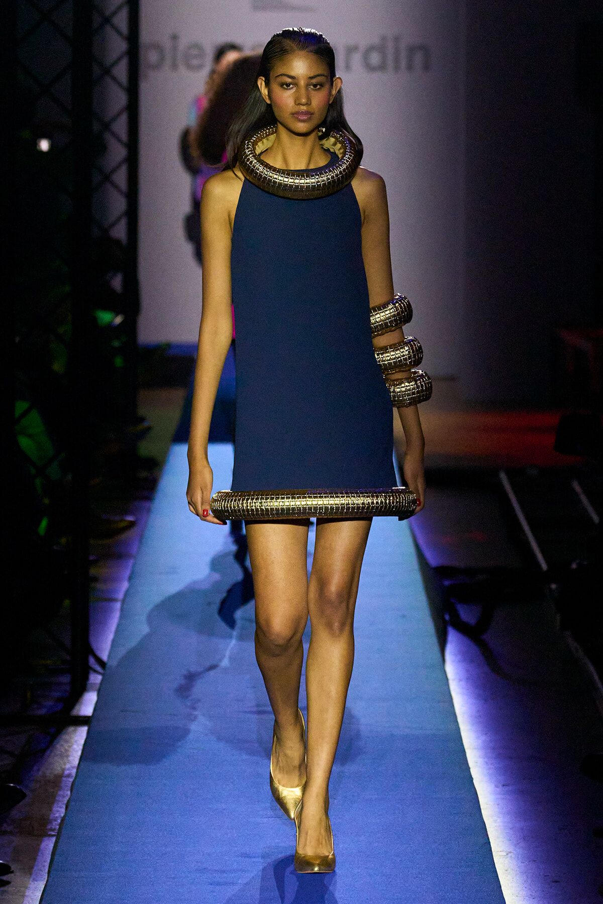 Model walking the runway in a navy blue sleeveless dress with a large gold coil neckpiece and matching gold bracelets/arm cuffs.