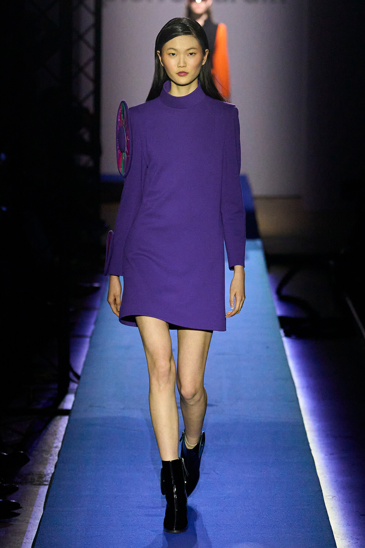 Model walks a fashion runway in a solid purple turtleneck mini dress with a circular shoulder accessory and black boots on a blue catwalk.