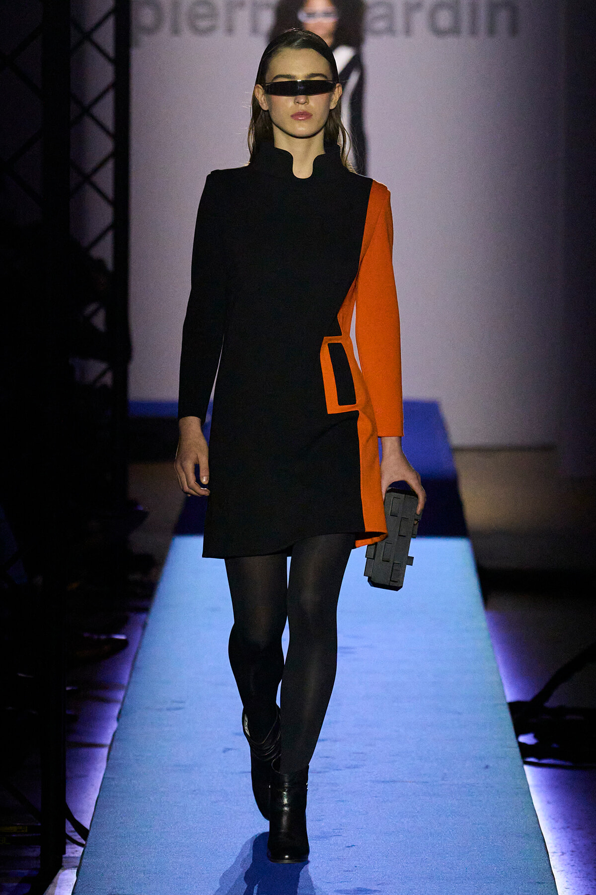 Model on a runway wearing a black and orange color-block dress with a futuristic black visor, black tights, and a gray geometric clutch.