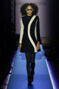 Model walking a fashion runway in a black dress with white sculpted stripes and futuristic sunglasses.