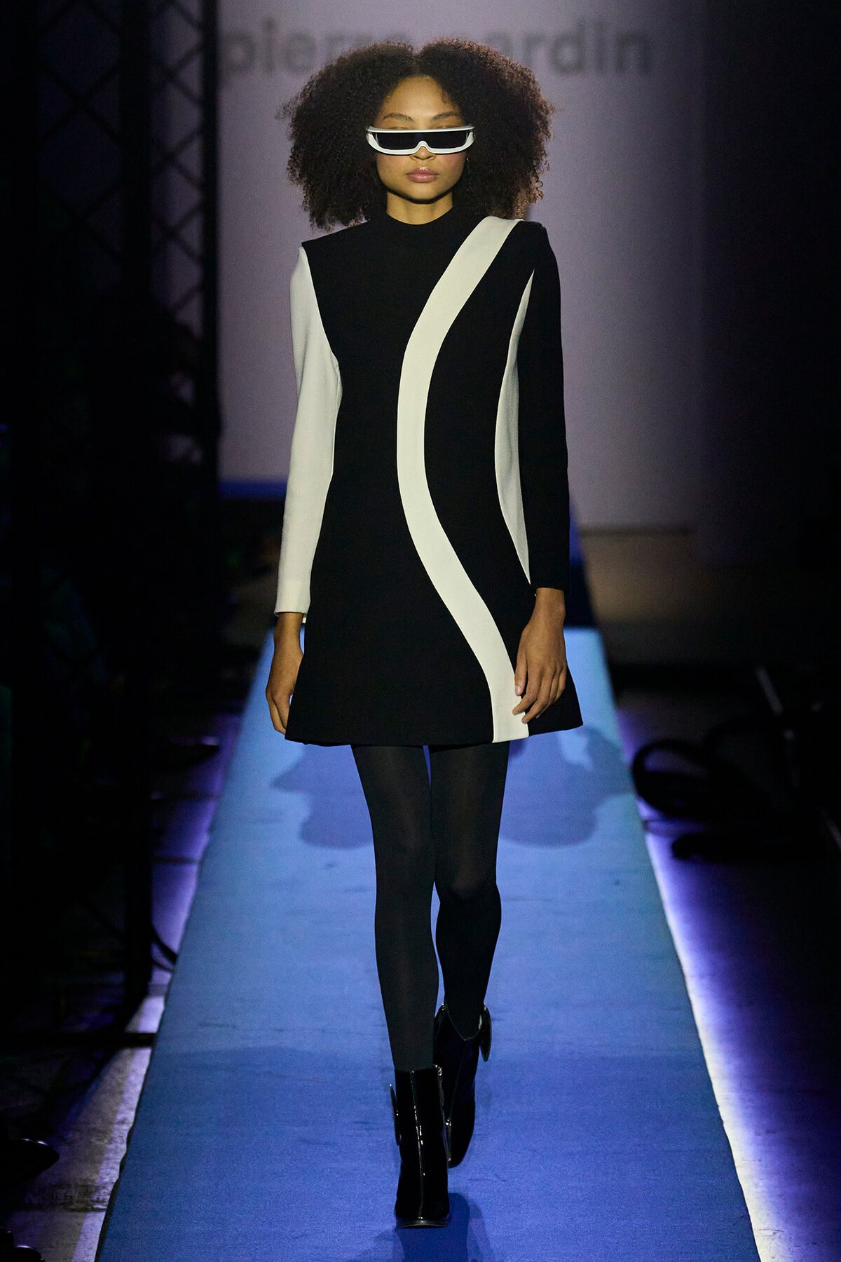 Model walking a fashion runway in a black dress with white sculpted stripes and futuristic sunglasses.