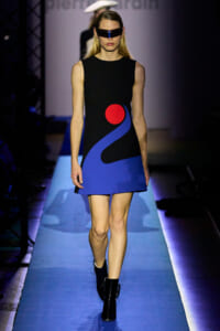 Model walks a runway in a black sleeveless dress with bold blue abstract shapes and a red circle; futuristic visor sunglasses add edge.
