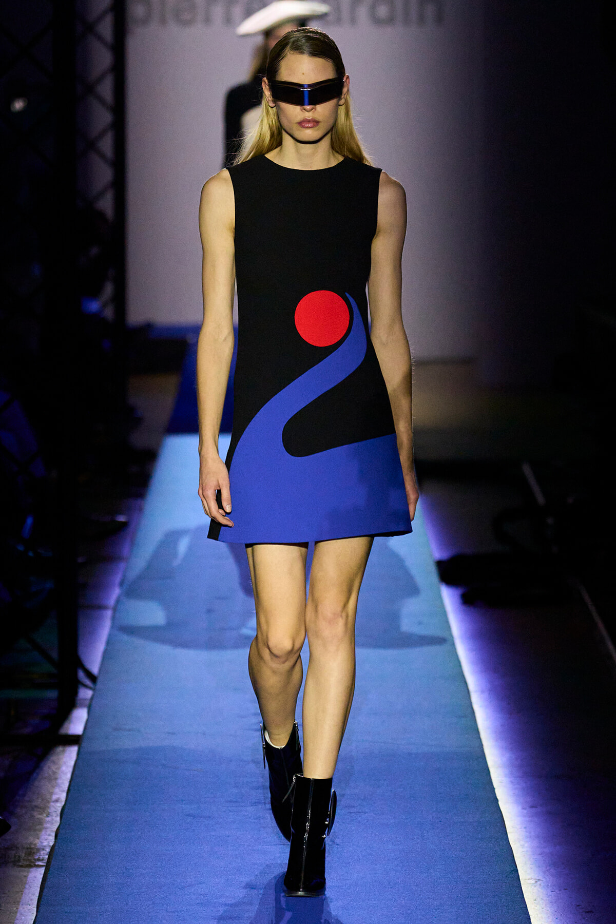 Model walks a runway in a black sleeveless dress with bold blue abstract shapes and a red circle; futuristic visor sunglasses add edge.