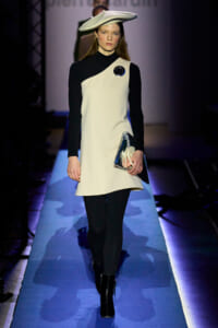 Model walking a runway in a black-and-white color-block dress with a matching white hat, holding a metallic clutch and wearing black boots.