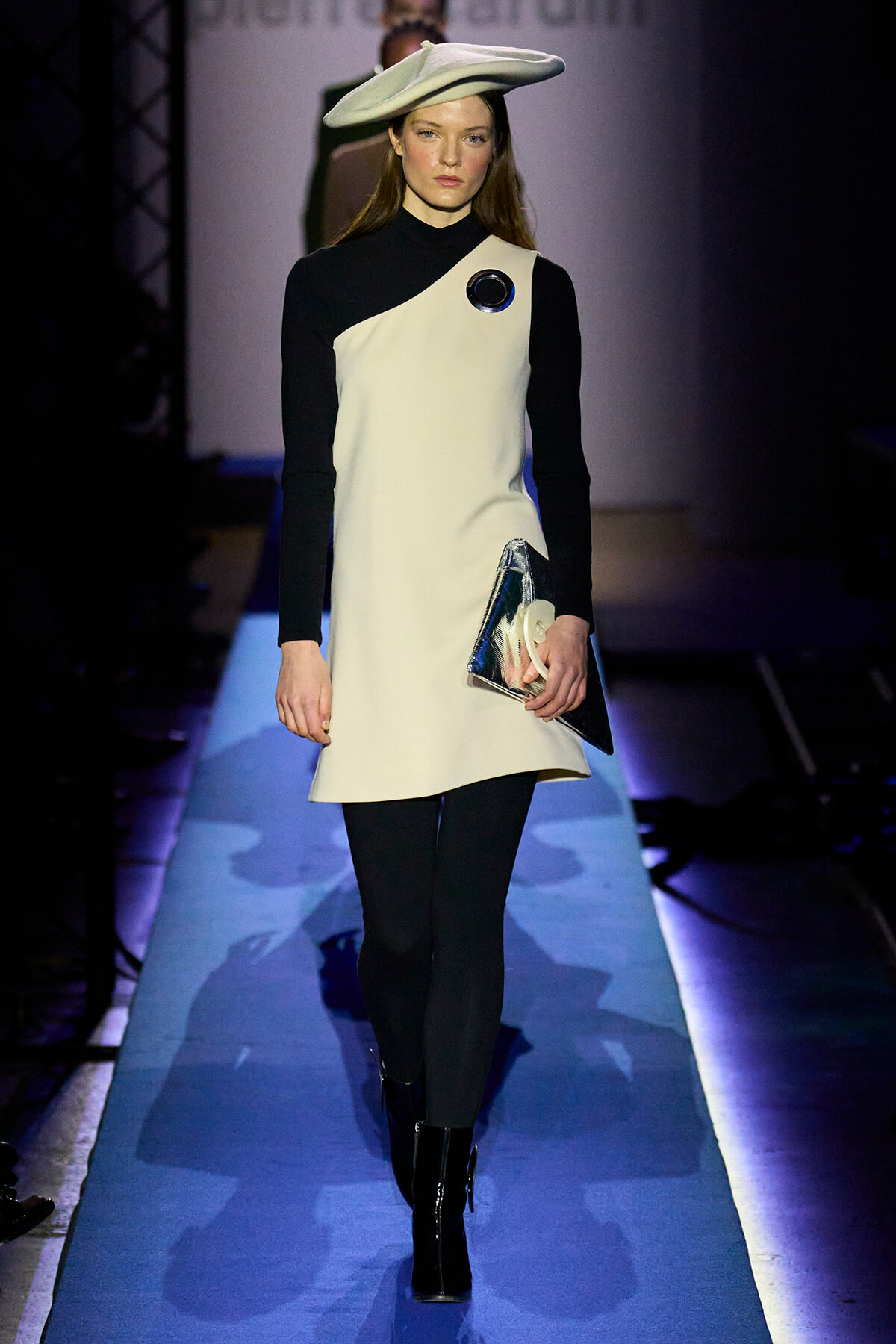 Model walking a runway in a black-and-white color-block dress with a matching white hat, holding a metallic clutch and wearing black boots.