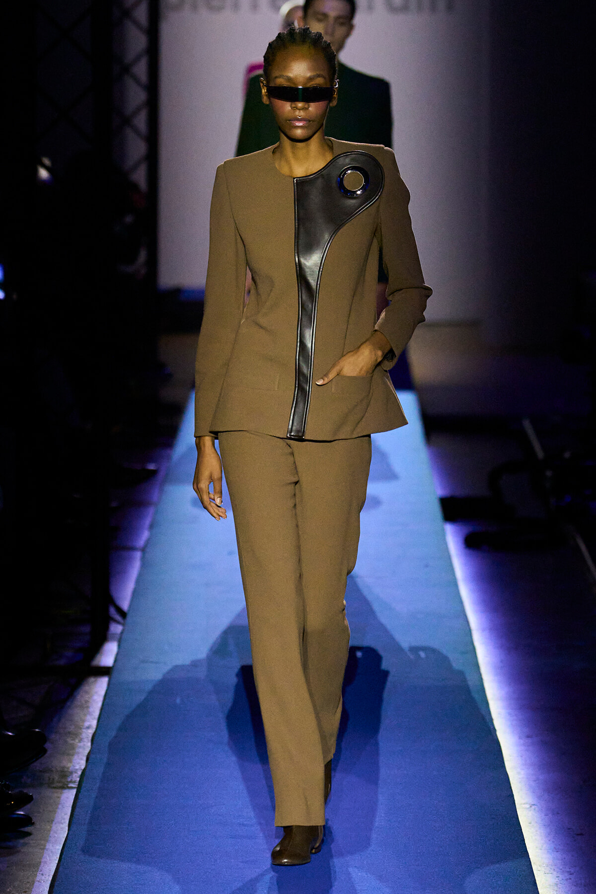 Model walks the runway in a camel pantsuit with a bold black leather curved panel and circular accent, wearing dark sunglasses.