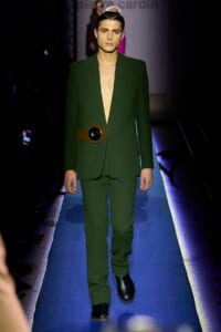 Male model walks the runway in a deep-plunge emerald green suit with a brown belt featuring a large circular buckle.