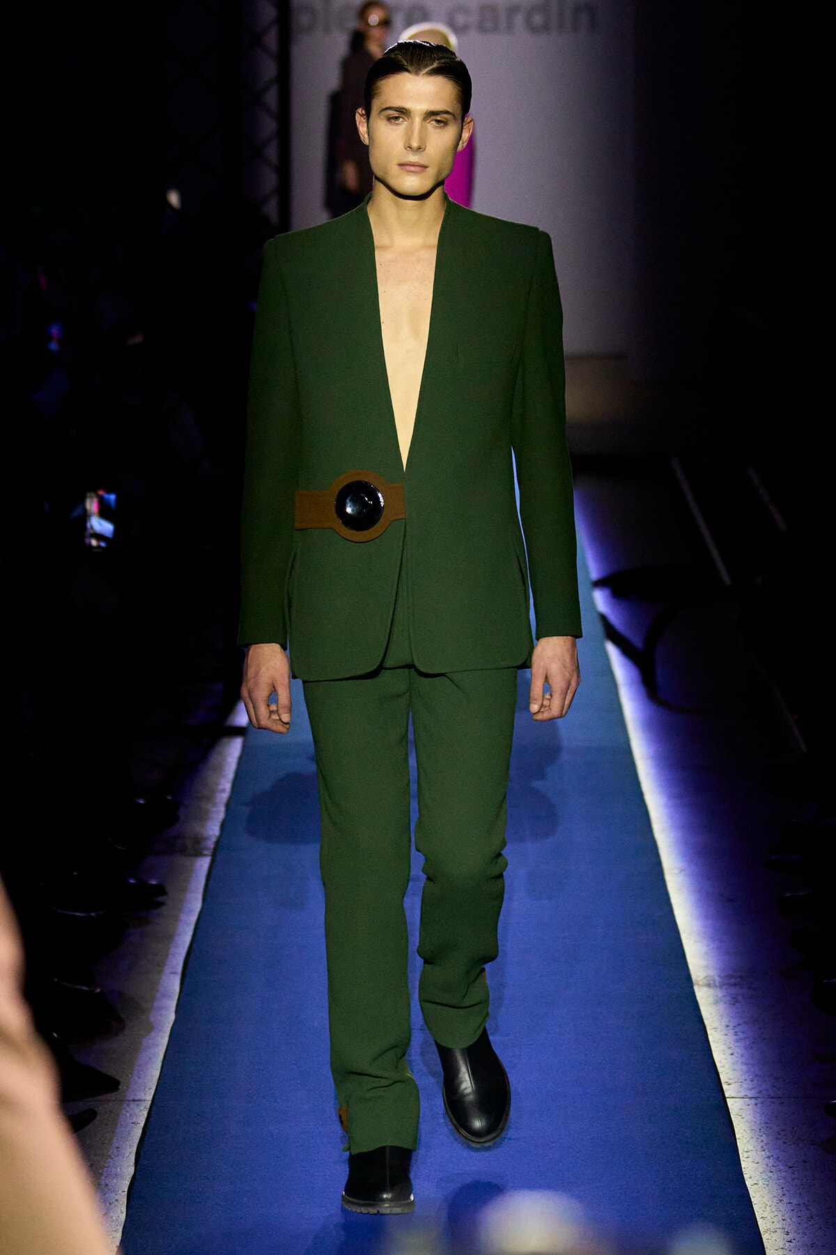 Male model walks the runway in a deep-plunge emerald green suit with a brown belt featuring a large circular buckle.