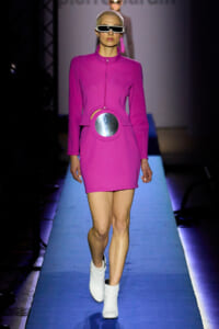 Model walking the runway in a bright pink mini dress with a circular metallic belt and futuristic white sunglasses on a blue-lit catwalk.