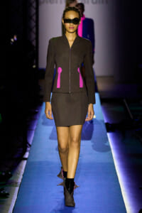 Model on a blue-lit runway wearing a dark brown zip-front jacket with pink pocket accents and a matching skirt; she wears black sunglasses MINIMAL