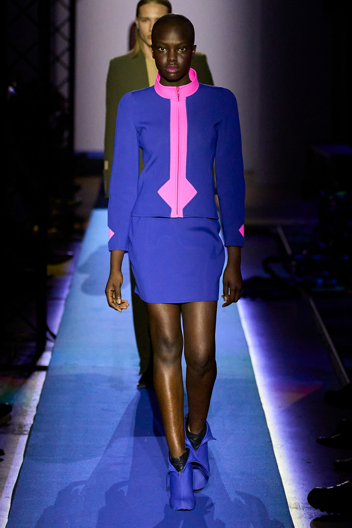 Model walks the runway in a cobalt blue dress with neon pink zipper and geometric pink accents at the collar and waist.
