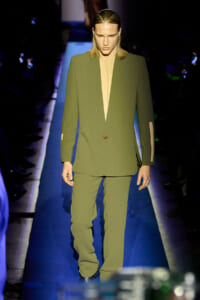 Male model on a runway wearing an olive-green suit with a beige deep-plunge shirt underneath.