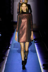 Model walking a runway in a metallic rose-gold and black long-sleeve mini dress with a high black collar, on a blue-lit catwalk.