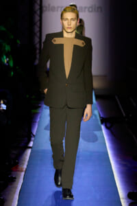 Male model walks a runway in a black suit with tan chest panels, hands in pockets, spotlight on him.