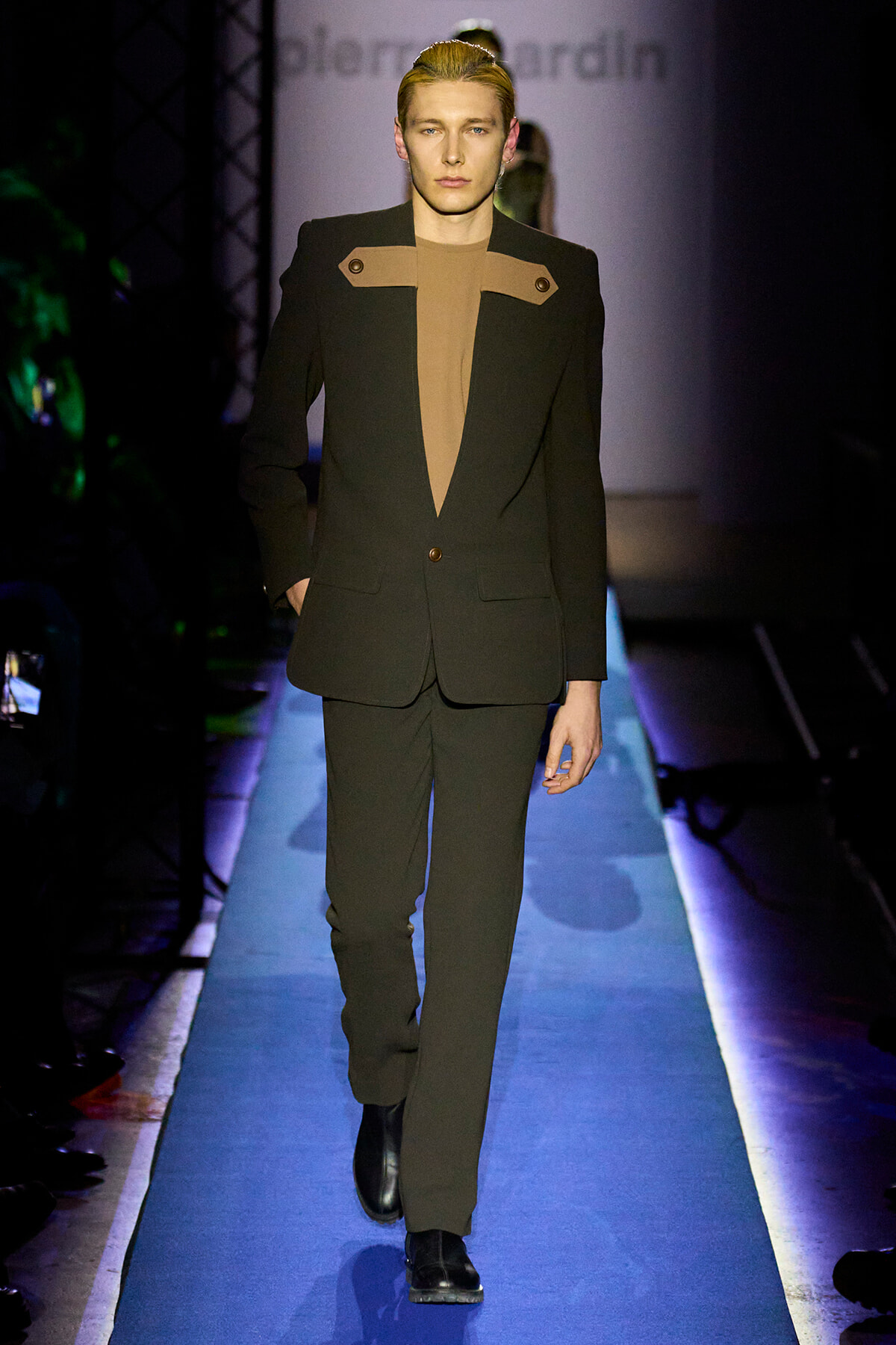 Male model walks a runway in a black suit with tan chest panels, hands in pockets, spotlight on him.