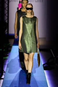Model walking a runway in a metallic green and black color-block dress with oversized sunglasses, carrying a brown handbag.