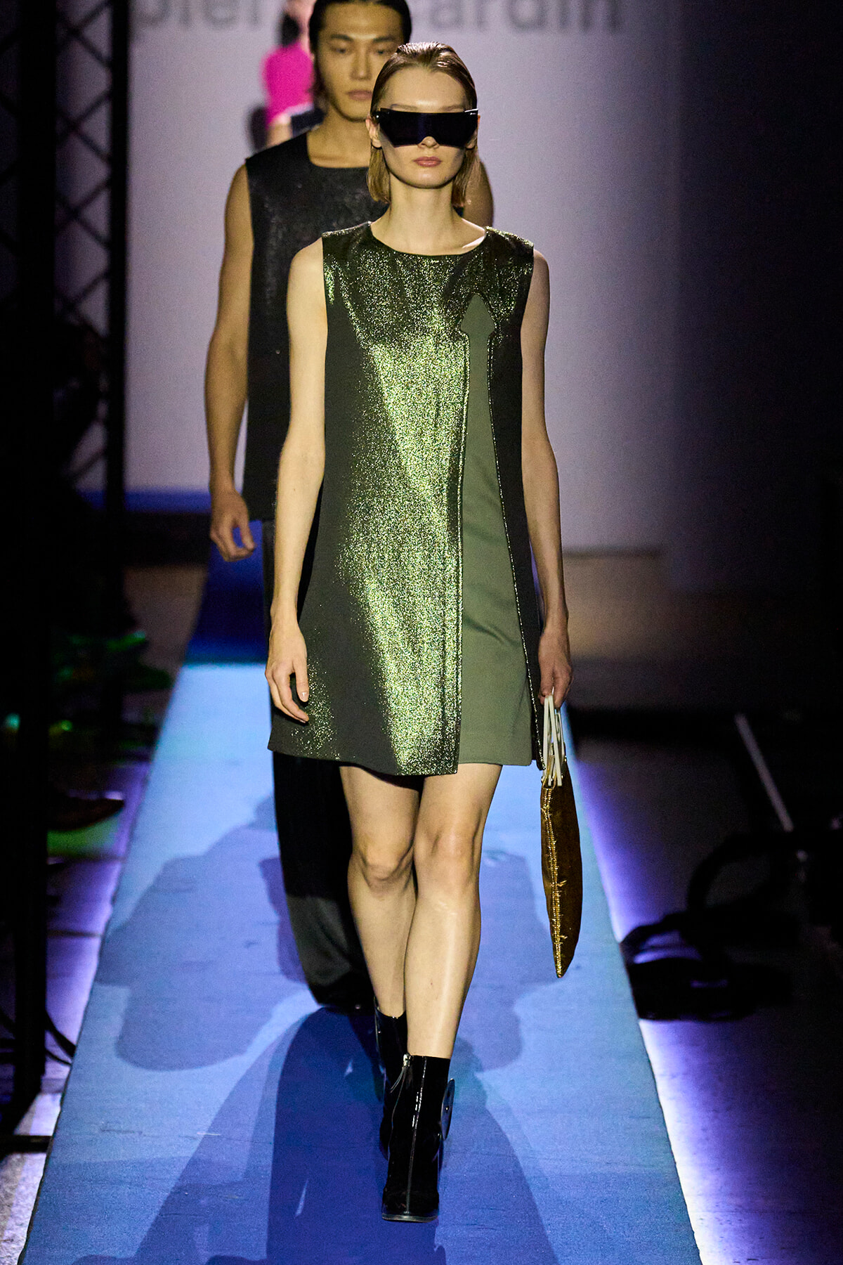 Model walking a runway in a metallic green and black color-block dress with oversized sunglasses, carrying a brown handbag.