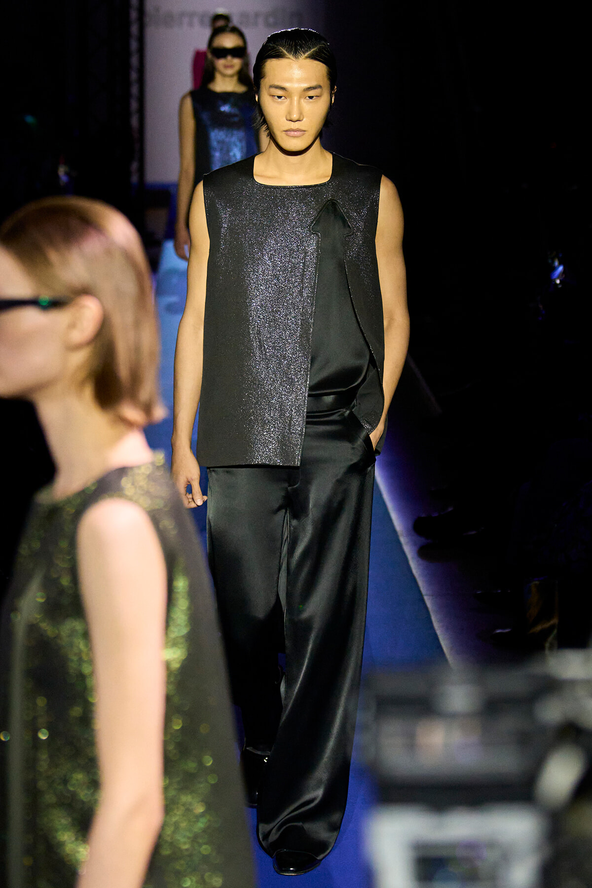 Model on a dark runway wearing a sleeveless glittery black vest over black satin pants; hair slicked back.