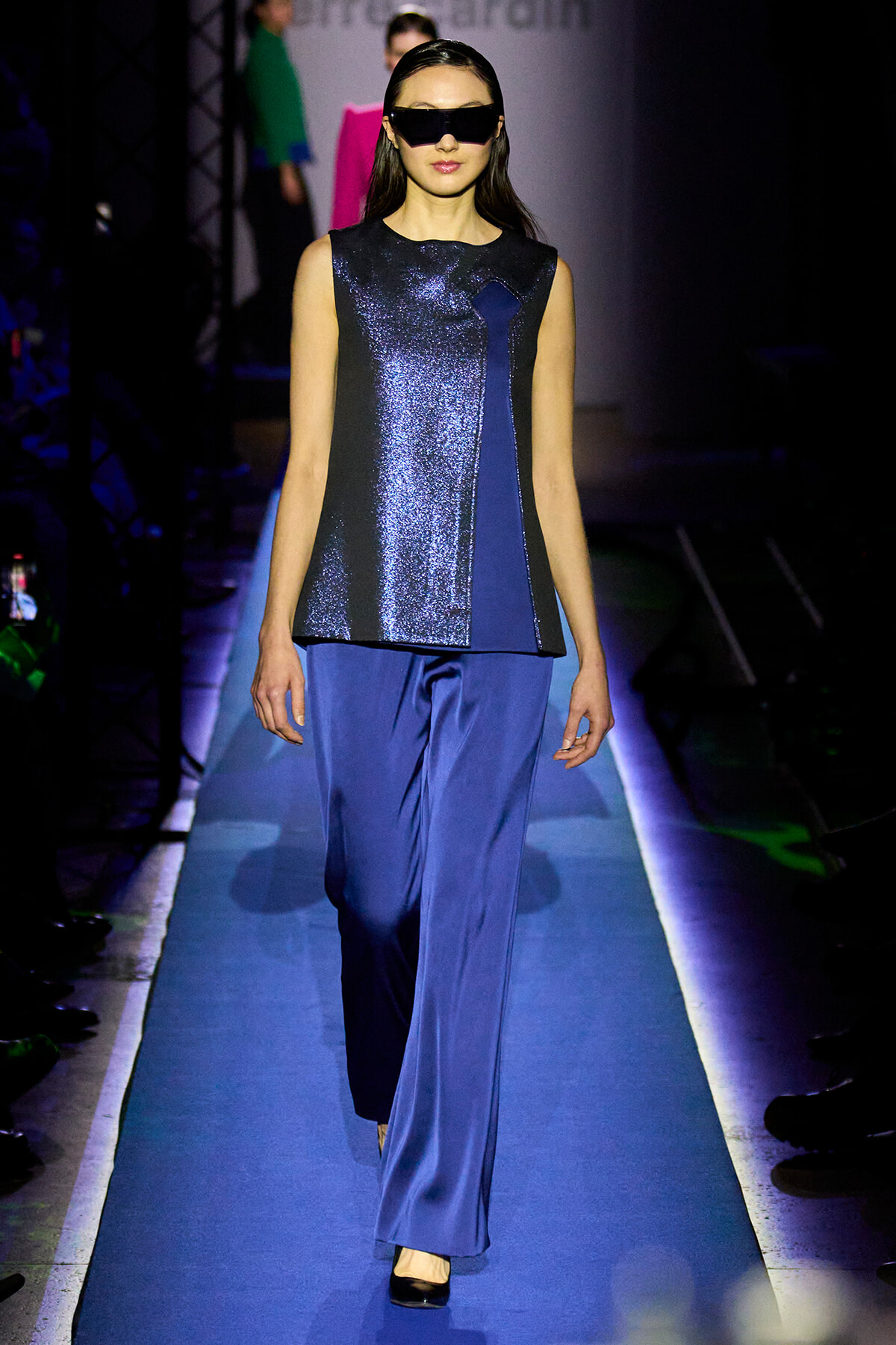 Model walks a blue metallic sleeveless top with matching wide-leg pants on a runway, wearing oversized black sunglasses.