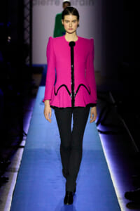Front-view of a fashion model walking the runway in a bright pink structured jacket with sculpted shoulders, black piping, and a peplum hem, paired with black pants and boots.