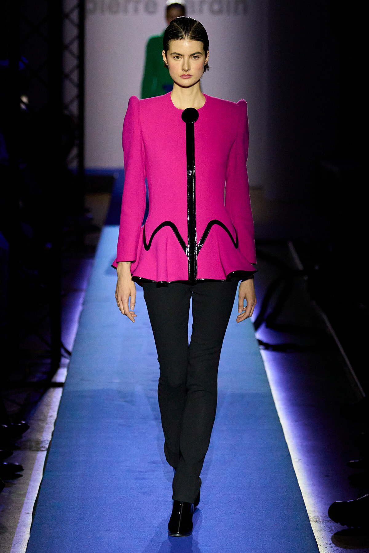 Front-view of a fashion model walking the runway in a bright pink structured jacket with sculpted shoulders, black piping, and a peplum hem, paired with black pants and boots.