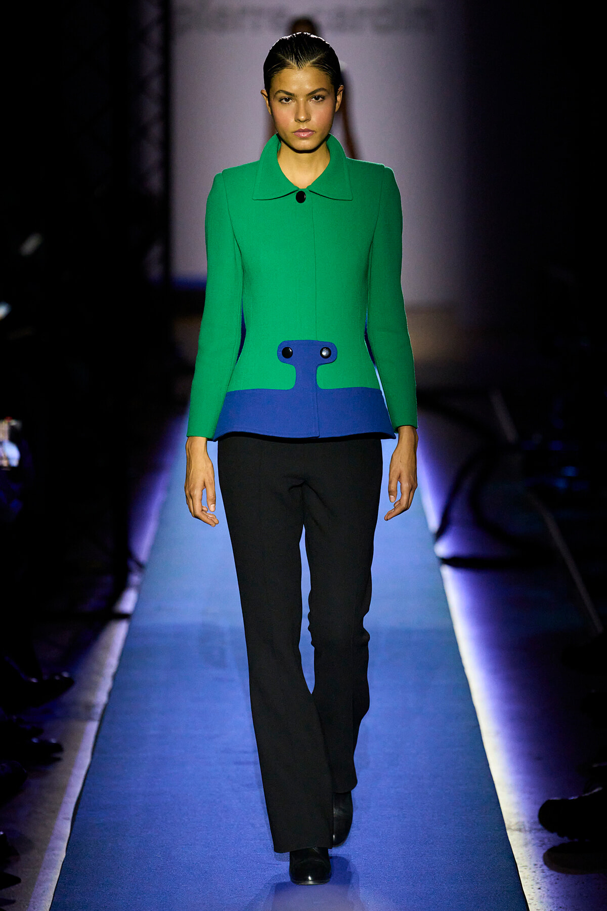 Runway model wearing a bright green coat with a blue waist panel and black pants, walking a blue-lit catwalk.