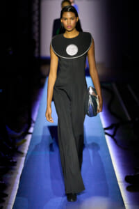 Model walks the runway in a black jumpsuit with a large circular silver neckline, carrying a metallic clutch.