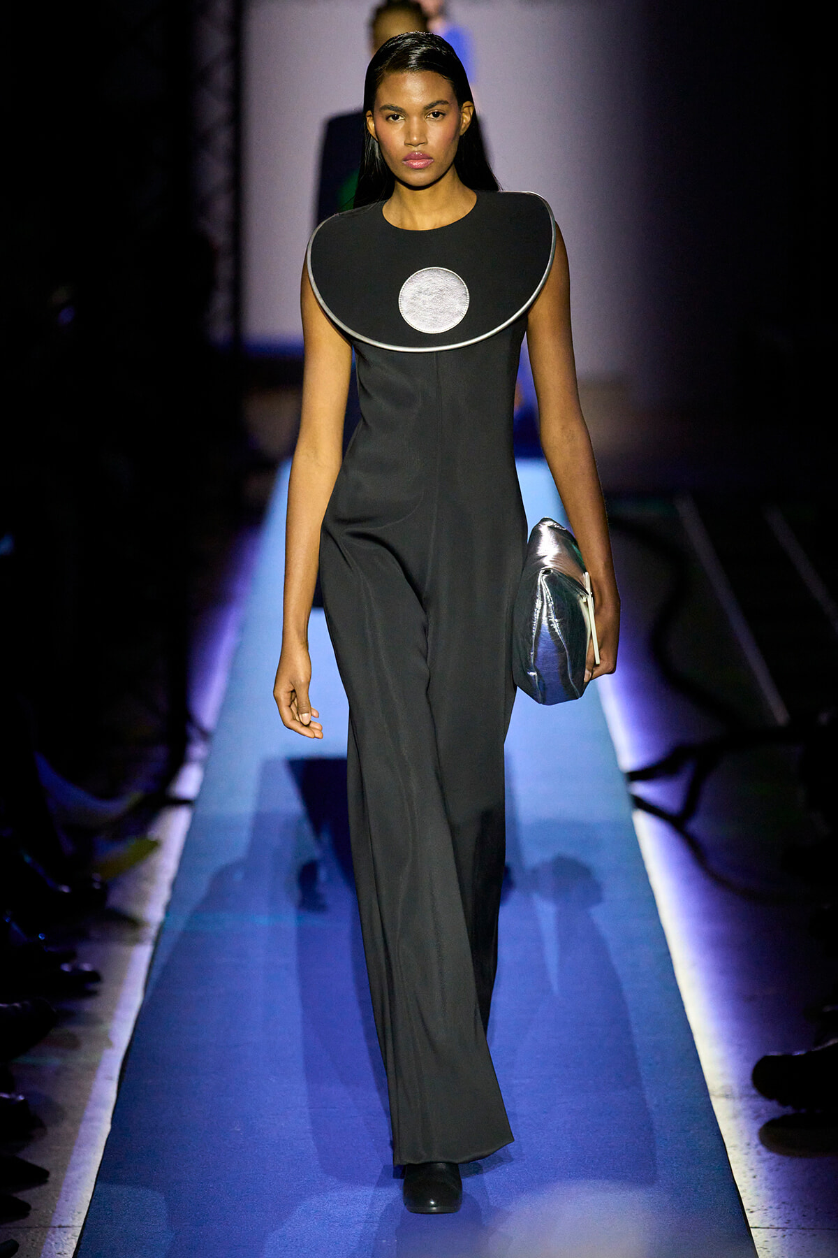 Model walks the runway in a black jumpsuit with a large circular silver neckline, carrying a metallic clutch.