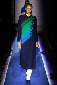 Model walking a runway in a navy coat with a bold diagonal rainbow panel and large black buttons; she holds a blue clutch.