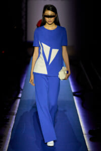 Model on a runway wearing a cobalt blue dress with white geometric panels, black visor sunglasses, and a white block clutch.