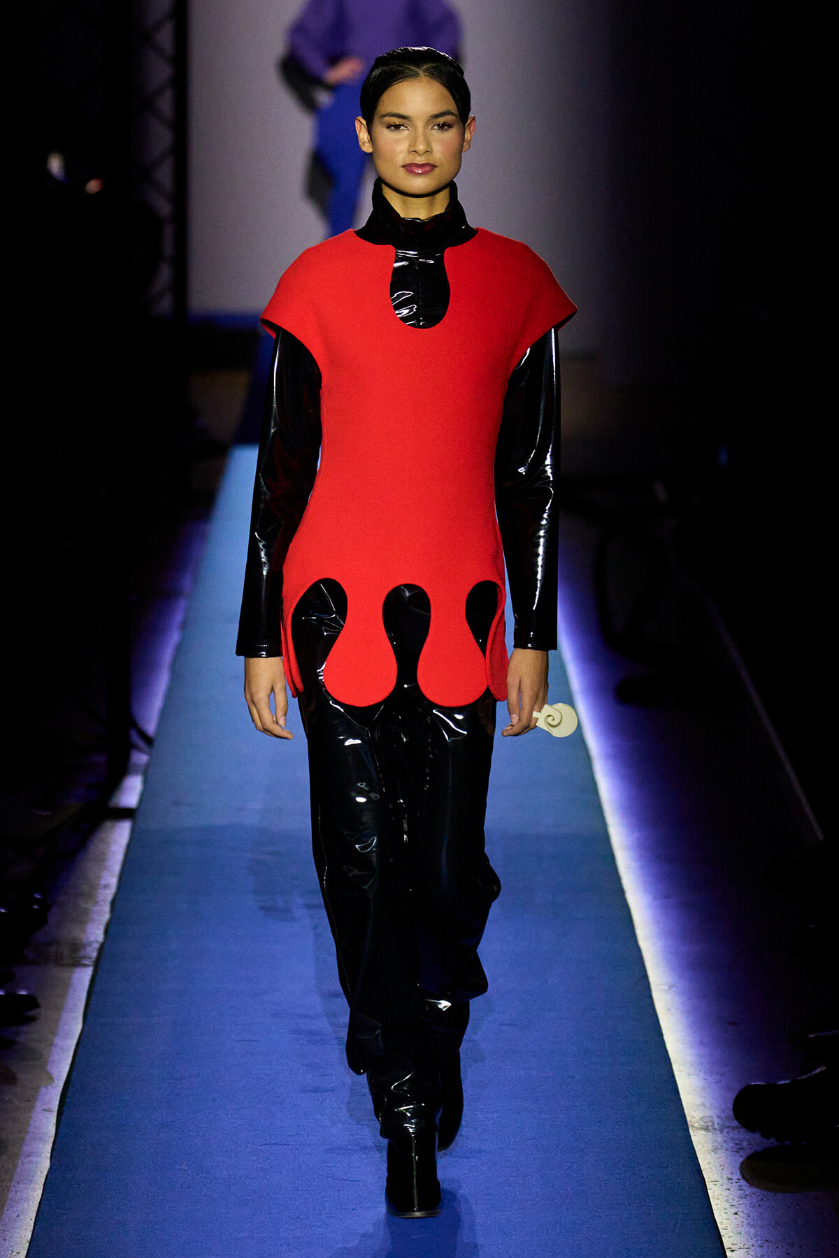 Model walking a runway in a striking red tunic with black patent sleeves and flame-like cutouts at the hem, black shiny pants, minimal makeup.