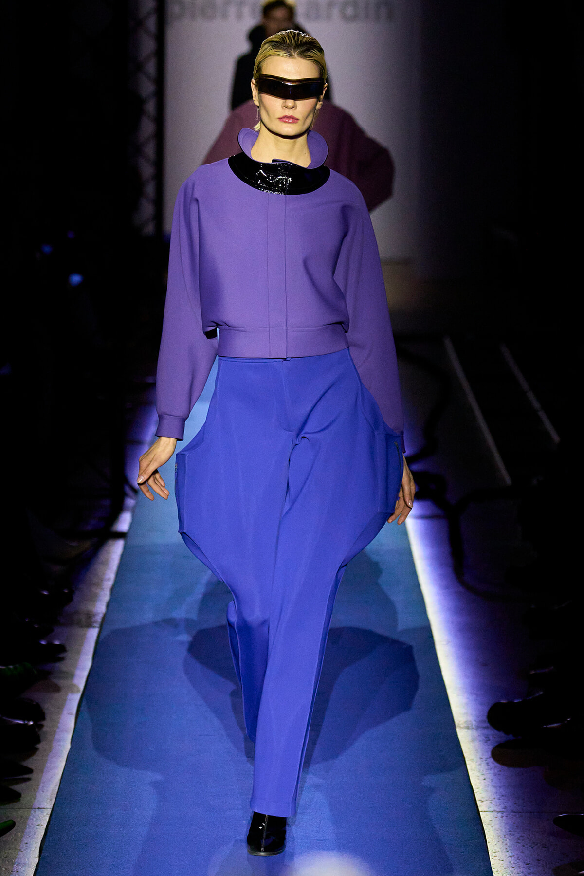 Model walks a futuristic runway in a purple top and cobalt-blue pants, wearing oversized black visor sunglasses and black ankle boots.