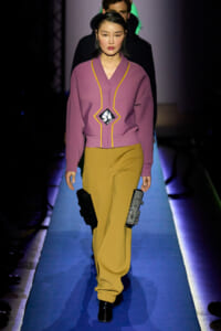 Model walks a runway in a mauve jacket with yellow piping and mustard wide-leg pants, holding black rectangular accessories at her sides.