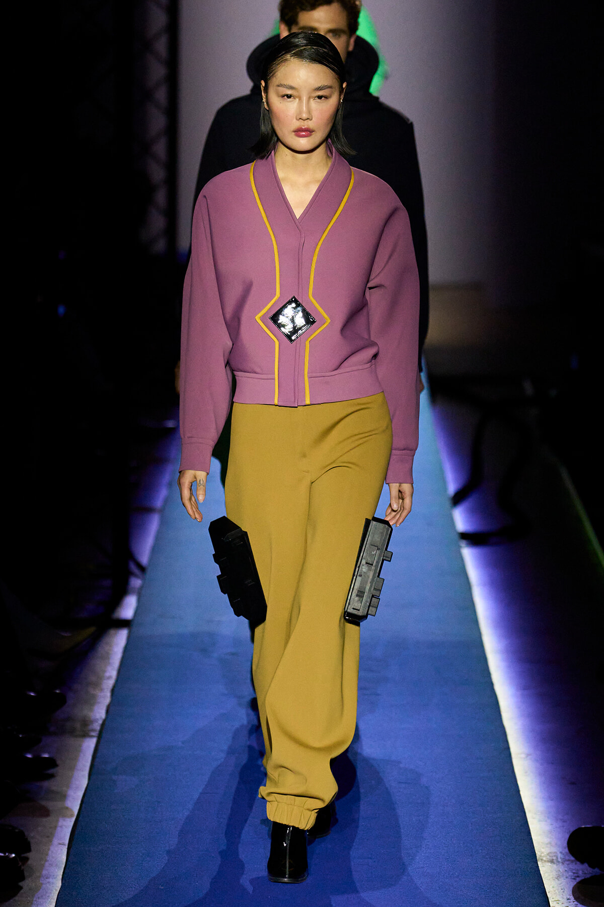 Model walks a runway in a mauve jacket with yellow piping and mustard wide-leg pants, holding black rectangular accessories at her sides.