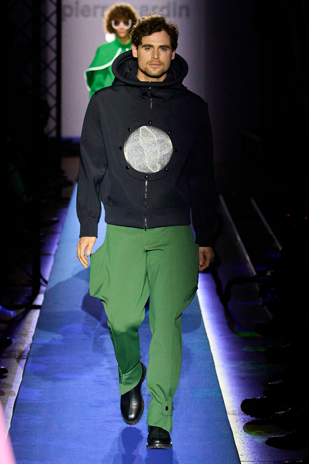 Male model on a runway wearing a black hoodie with a large glittery silver circle on the chest and bright green pants, walking forward confidently.