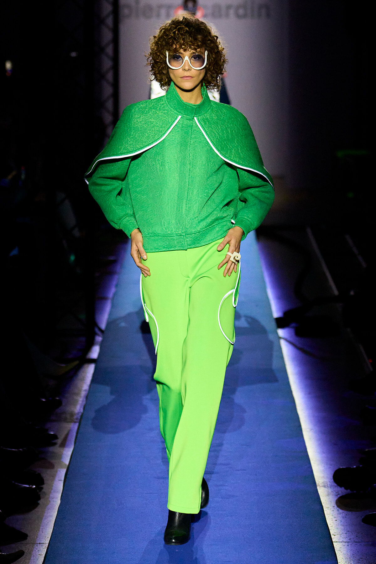 Model walking a fashion runway in a bright green cape-like jacket and matching lime pants with white piping, black ankle boots, and oversized white sunglasses.
